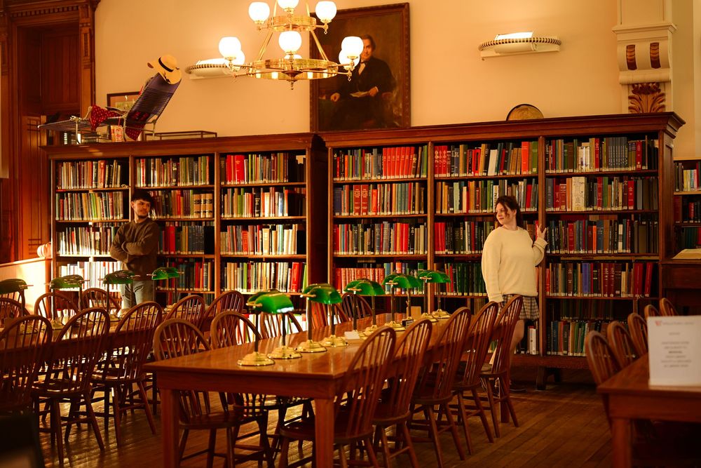 A warm, cozy library interior with wooden tables and chairs illuminated by soft lighting and lined with bookshelves.