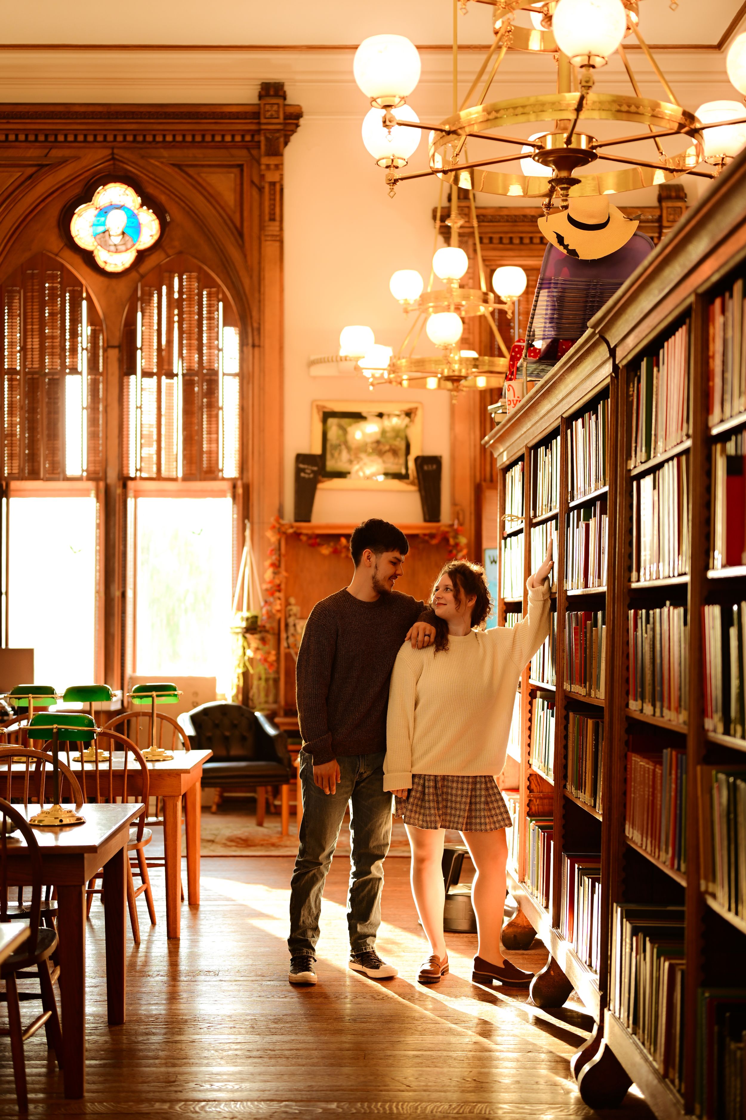 Warm sunlight streams through gothic windows in a historic library with wooden bookshelves and vintage chandeliers.