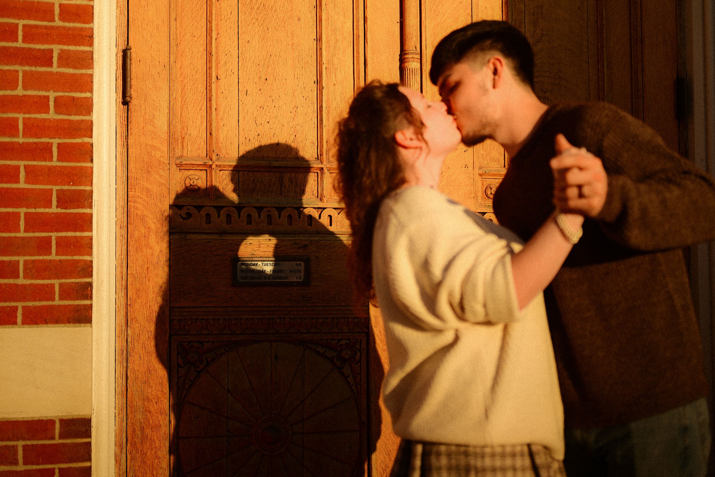 A couple embraces in a romantic moment against a brick wall with warm lighting.