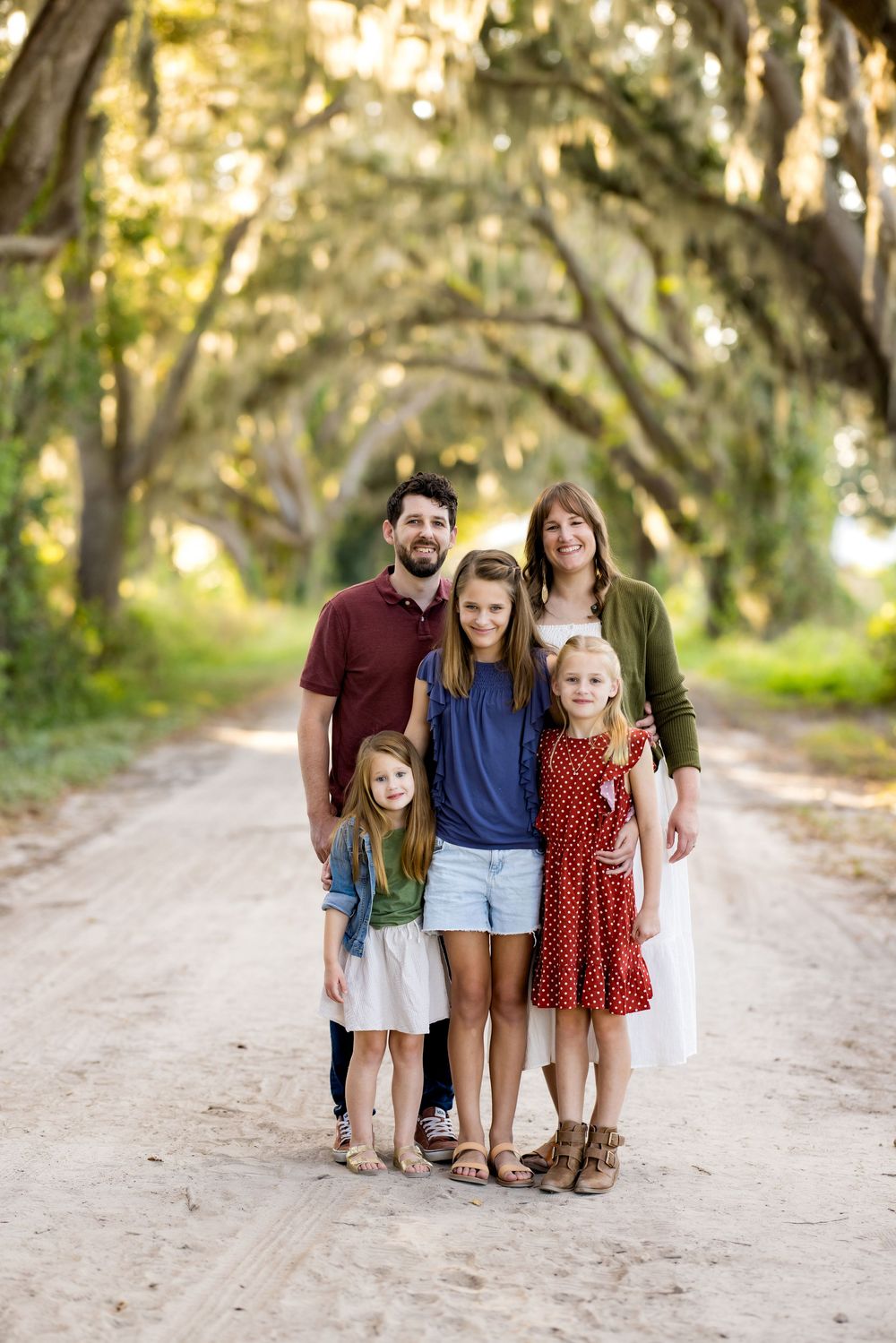 A family stands together on a sunlit dirt path surrounded by oak trees in a natural outdoor setting.