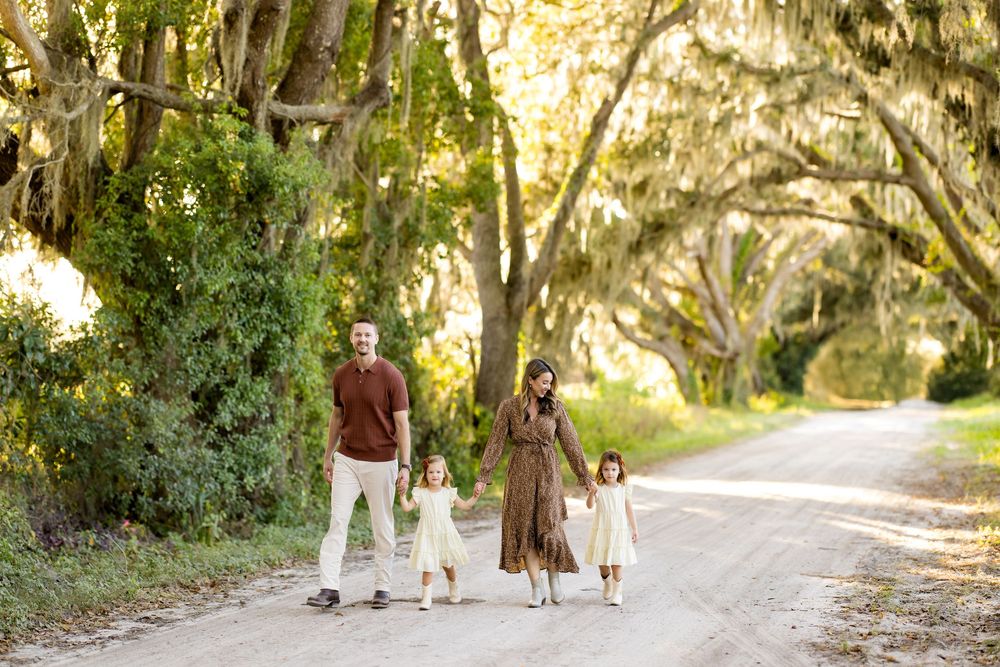 A dog walks with people on a sunlit tree-lined dirt path during a peaceful afternoon stroll.