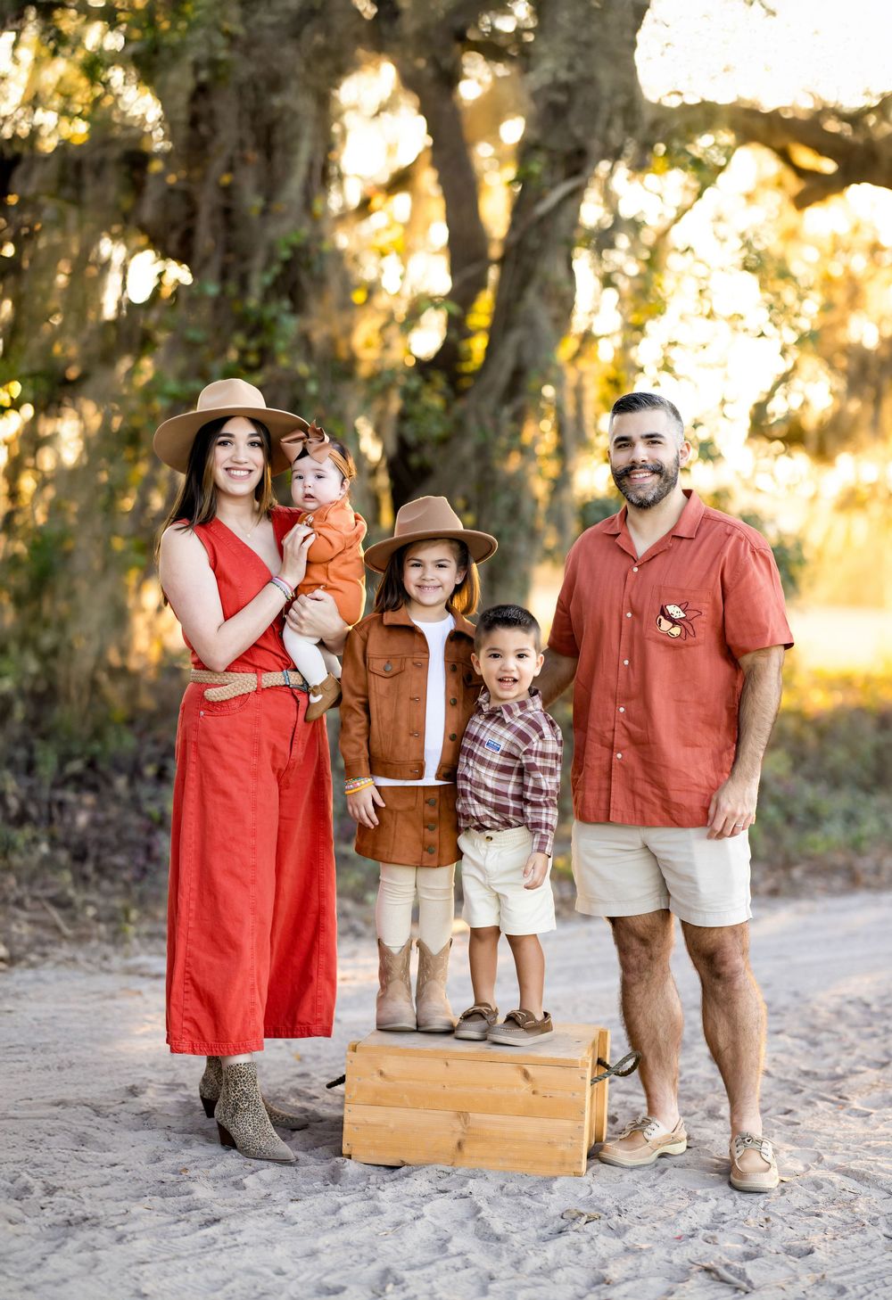 A family in coordinating coral and brown outfits poses together on a dirt path during golden hour sunset.