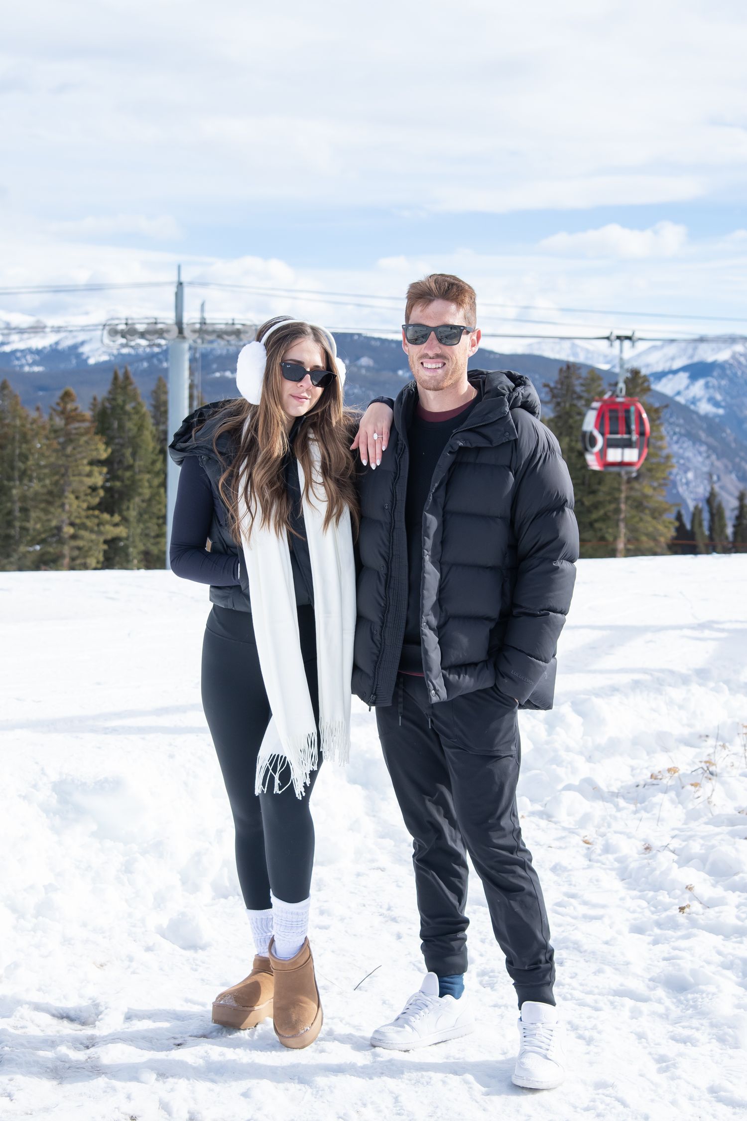Cute and chic couple stands together on top of Aspen mountain with a ski gondola in the background after a surprise proposal.
