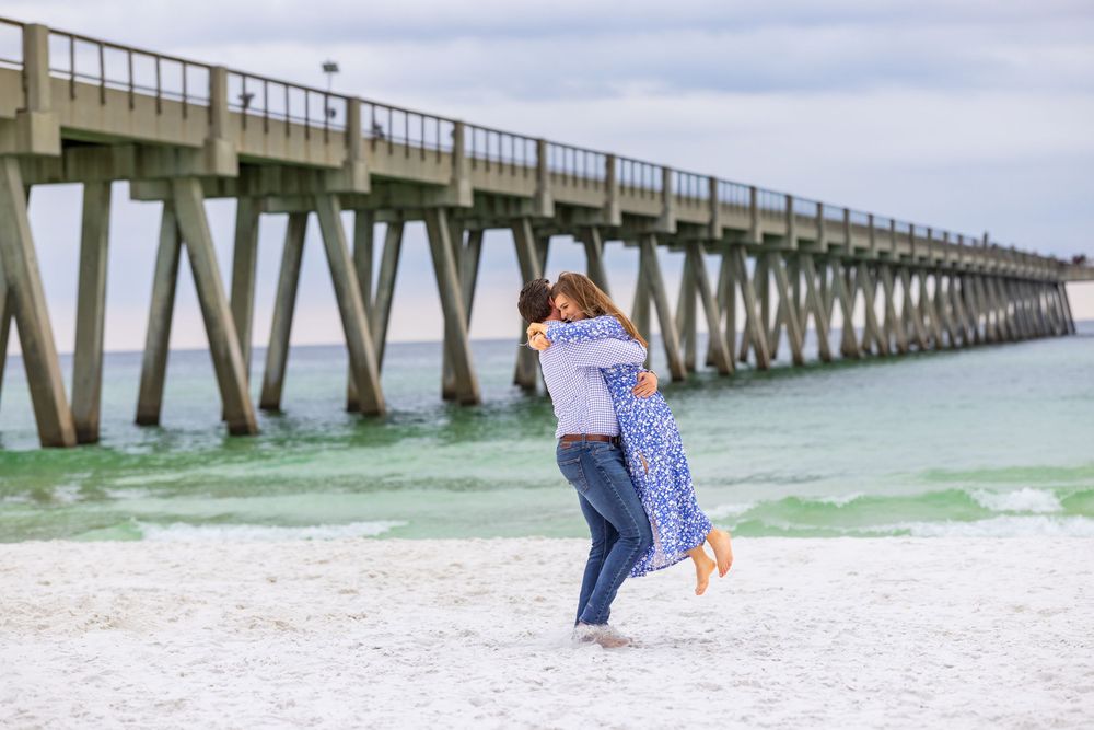 A couple embraces on a white sandy beach with a long wooden fishing pier stretching into turquoise waters.