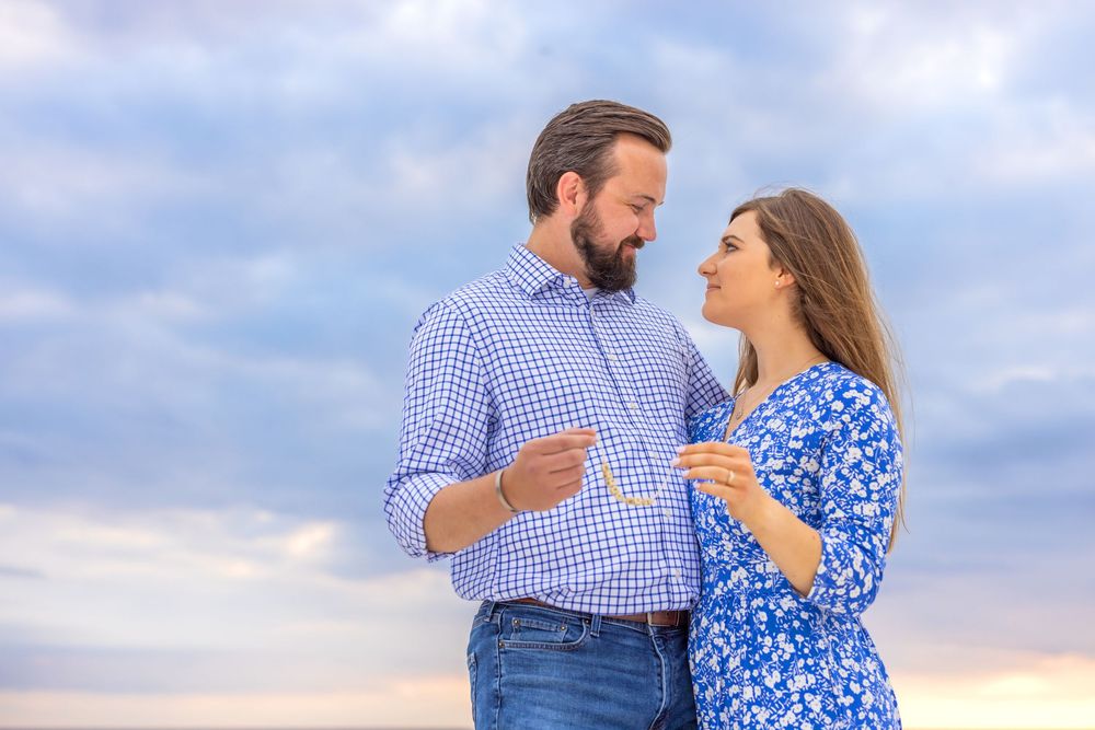 A couple in blue and white outfits poses for romantic engagement photos at sunset against a cloudy sky.