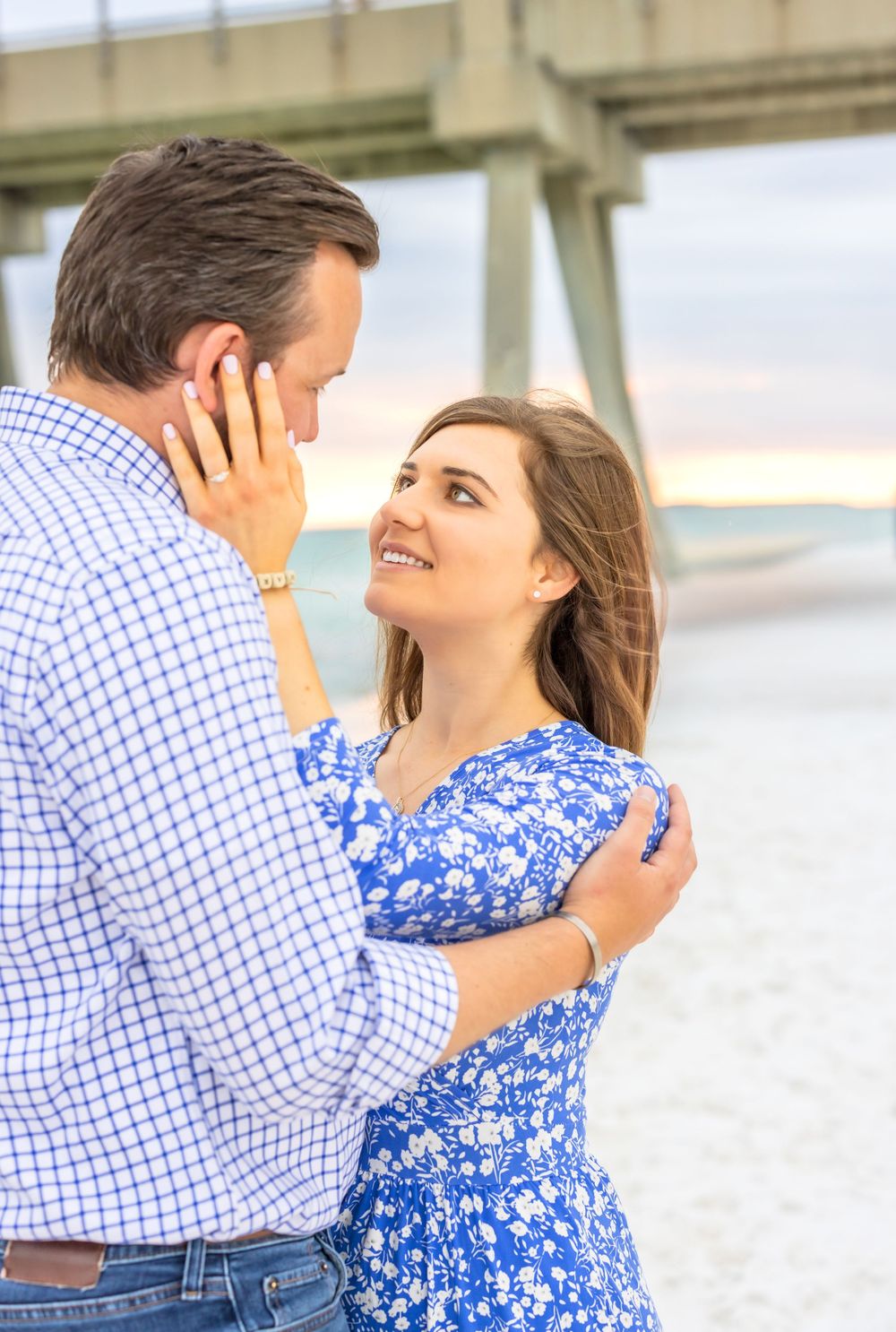 Couple in a blue floral dress and checkered shirt embrace lovingly on a bright sunny day at the beach.