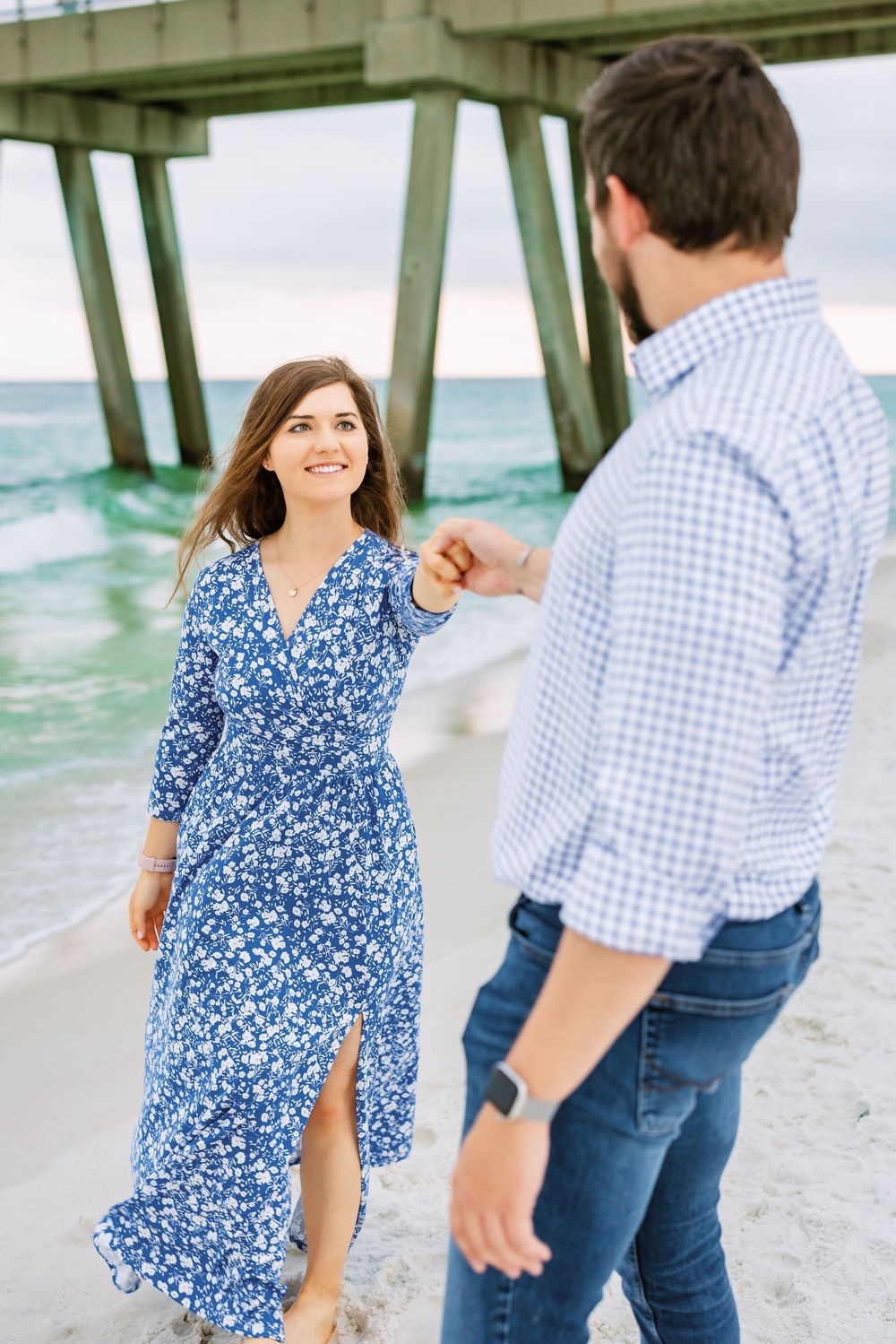 Couple walking and laughing together on a beach under a wooden pier in casual blue and white outfits.
