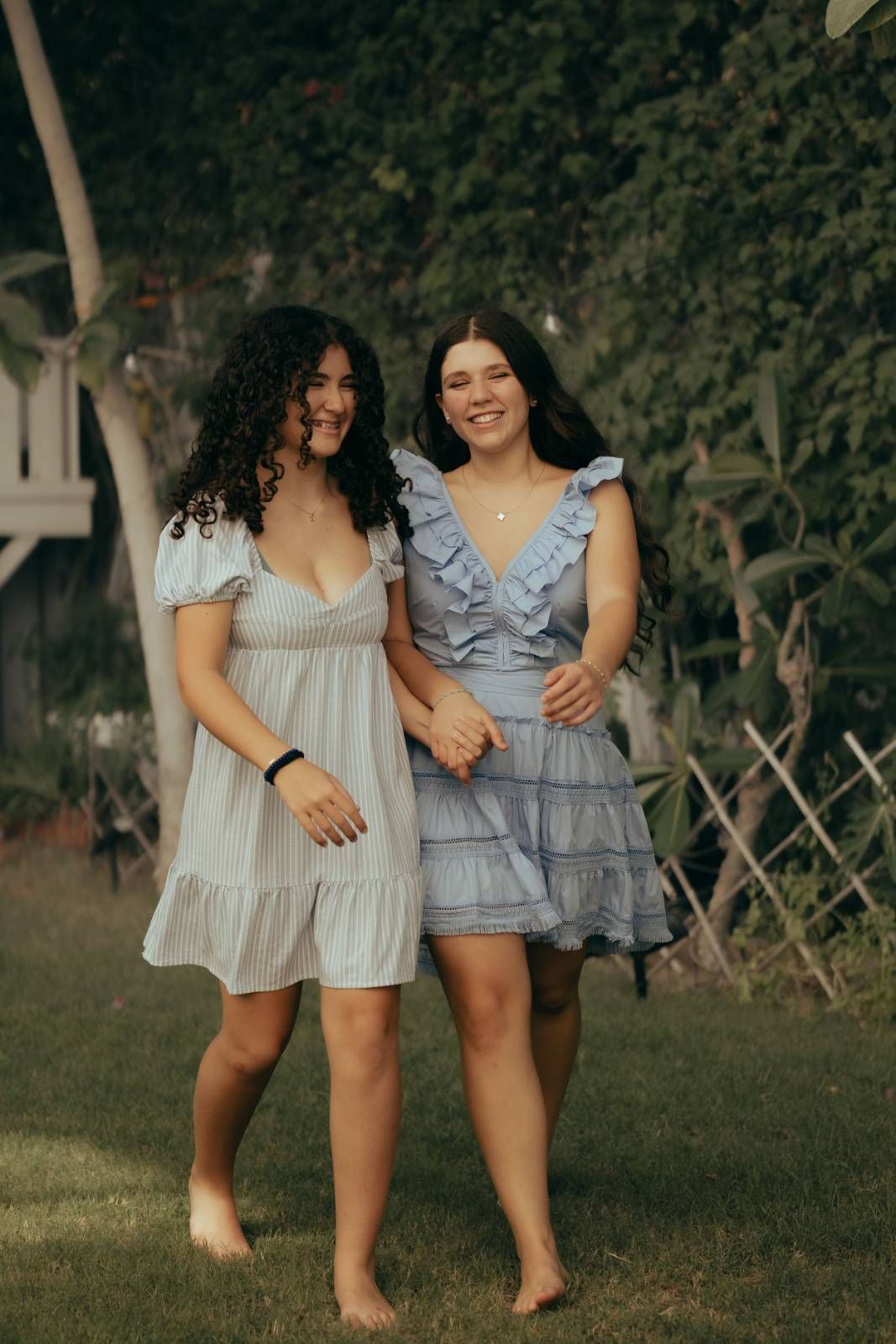 Two teenage girls in beige dresses posing together outdoors in green garden setting