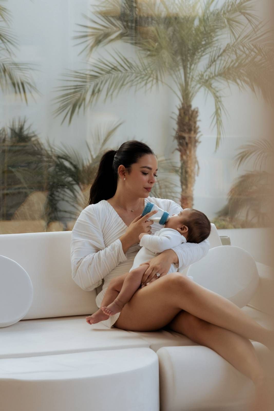 Woman in white sleeveless top holding baby near palm tree indoors