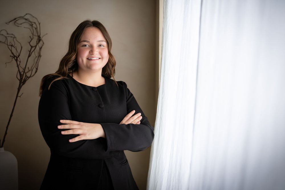 Professional headshot portrait against white backdrop in black sweater with crossed arms and warm lighting.