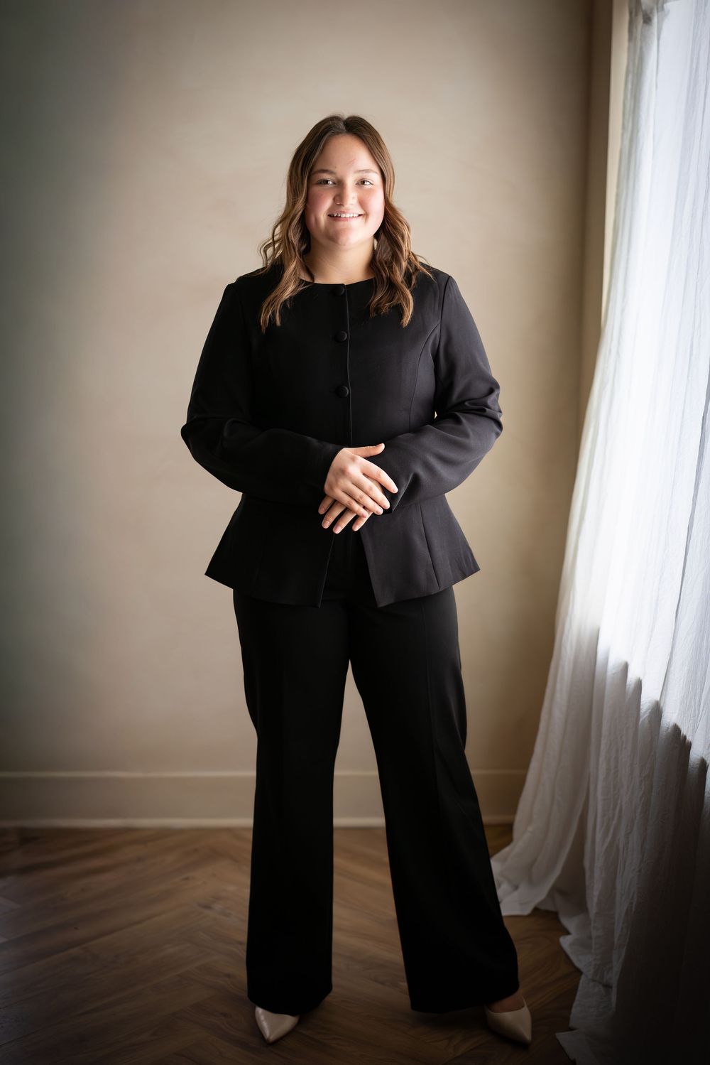 A person in a black business suit poses against a light wall with sheer curtains in a series of dynamic shots.