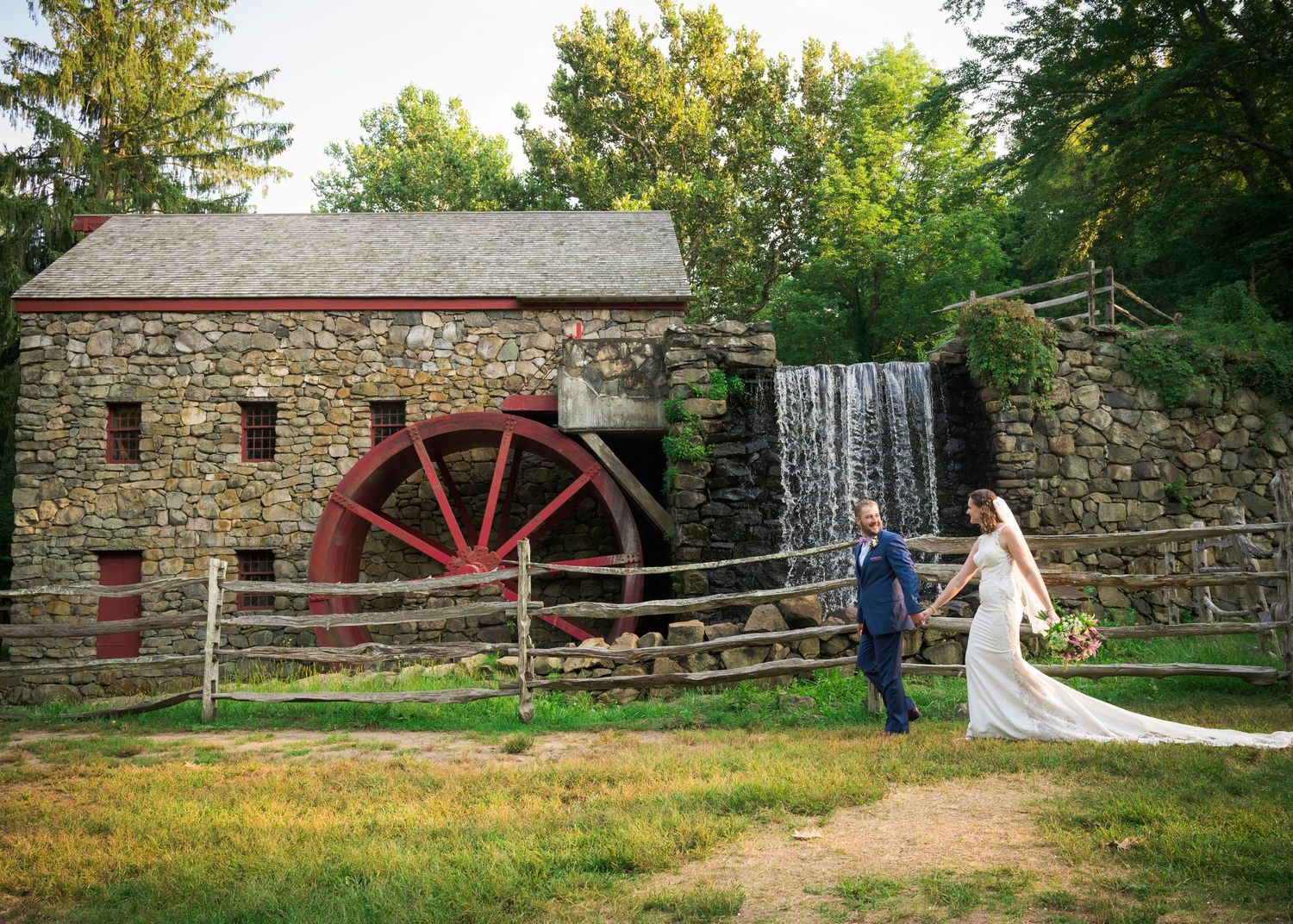 Historic stone mill with red waterwheel and cascading waterfall surrounded by lush green trees and landscaping.