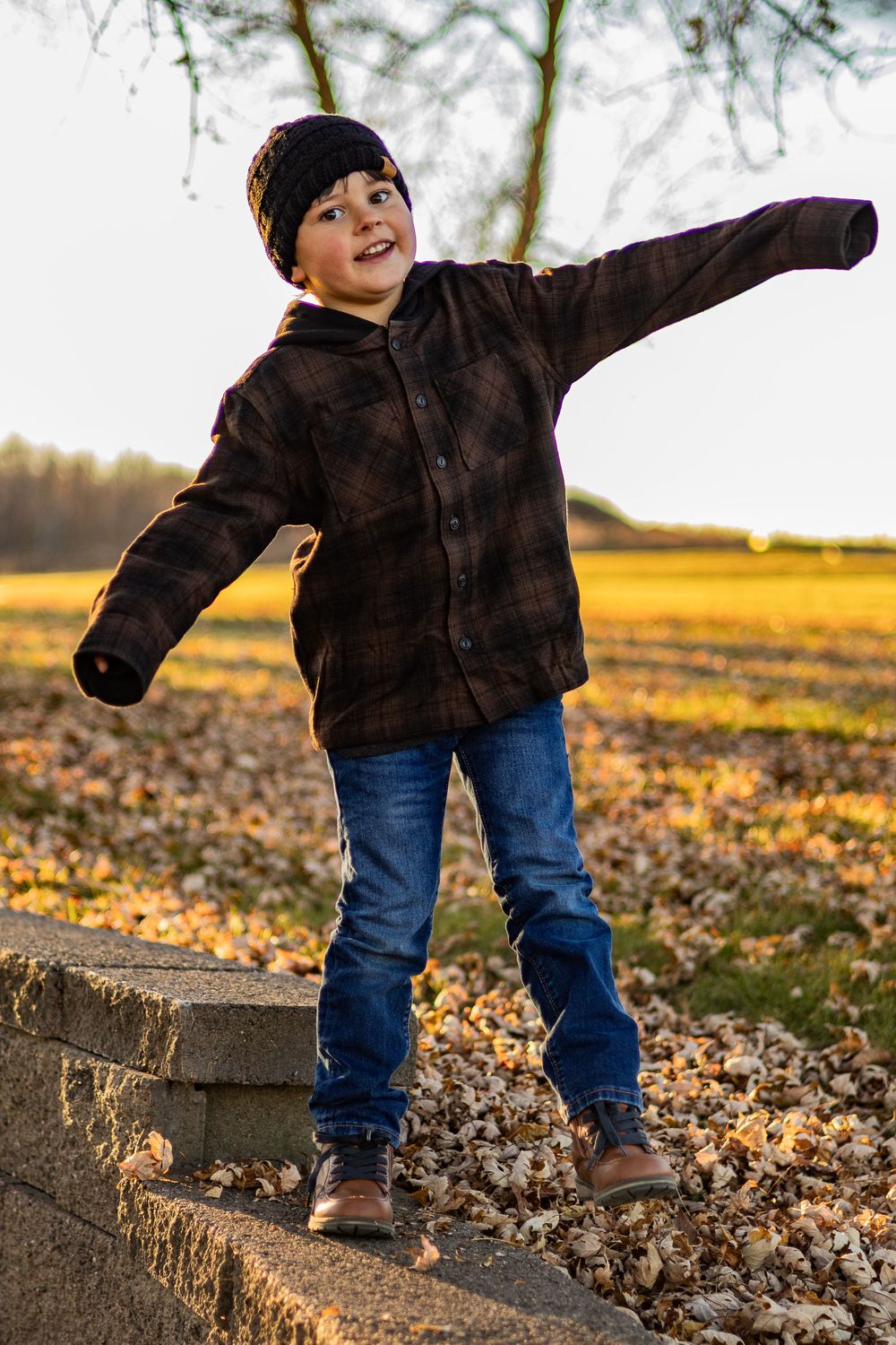 Child in brown jacket and jeans plays on concrete wall surrounded by fallen leaves at sunset.