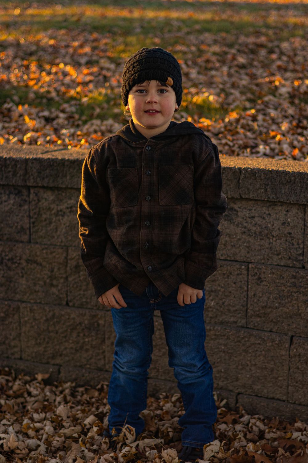Child plays near concrete wall during sunset with autumn leaves scattered on ground.