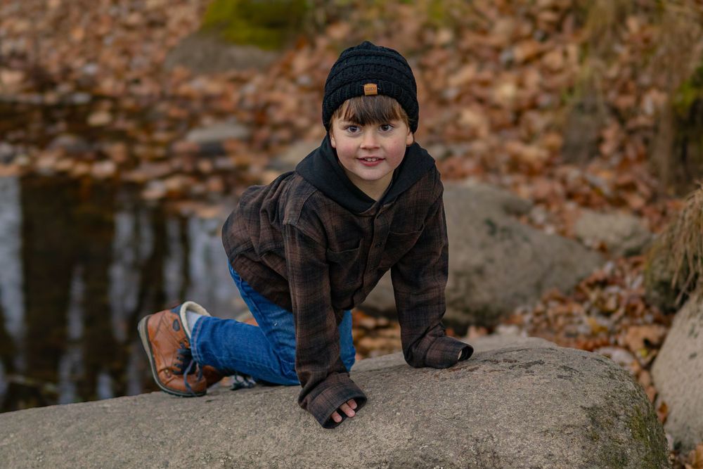 Person in winter clothing climbs on large rock near water surrounded by fallen leaves.