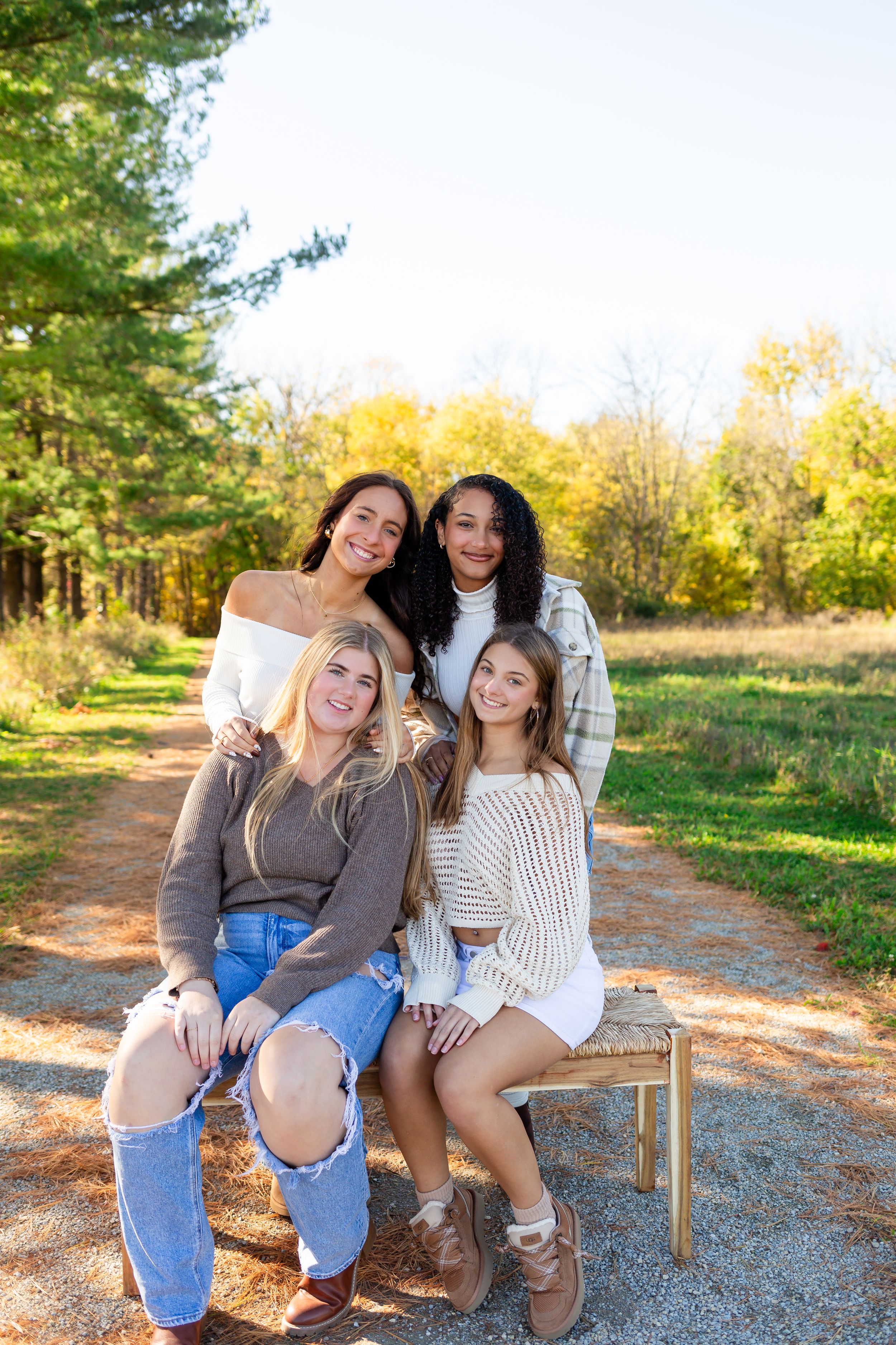 Four friends sit together on a wooden bench along a dirt path surrounded by autumn trees and golden sunlight.