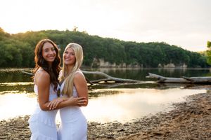 Two people in white dresses stand together at sunset by a serene lake with trees in the background.