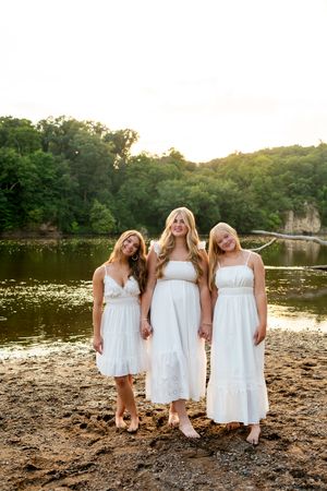 Three friends in white dresses sit together on a rocky beach at sunset near a tranquil lake with trees in the background.
