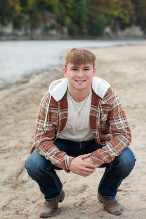 Casual portrait of someone in a plaid flannel jacket and jeans crouching on a beach with water in the background.