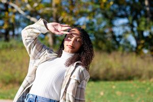 A person with curly hair wearing a white sweater and jeans shields their eyes from sunlight in an outdoor setting.