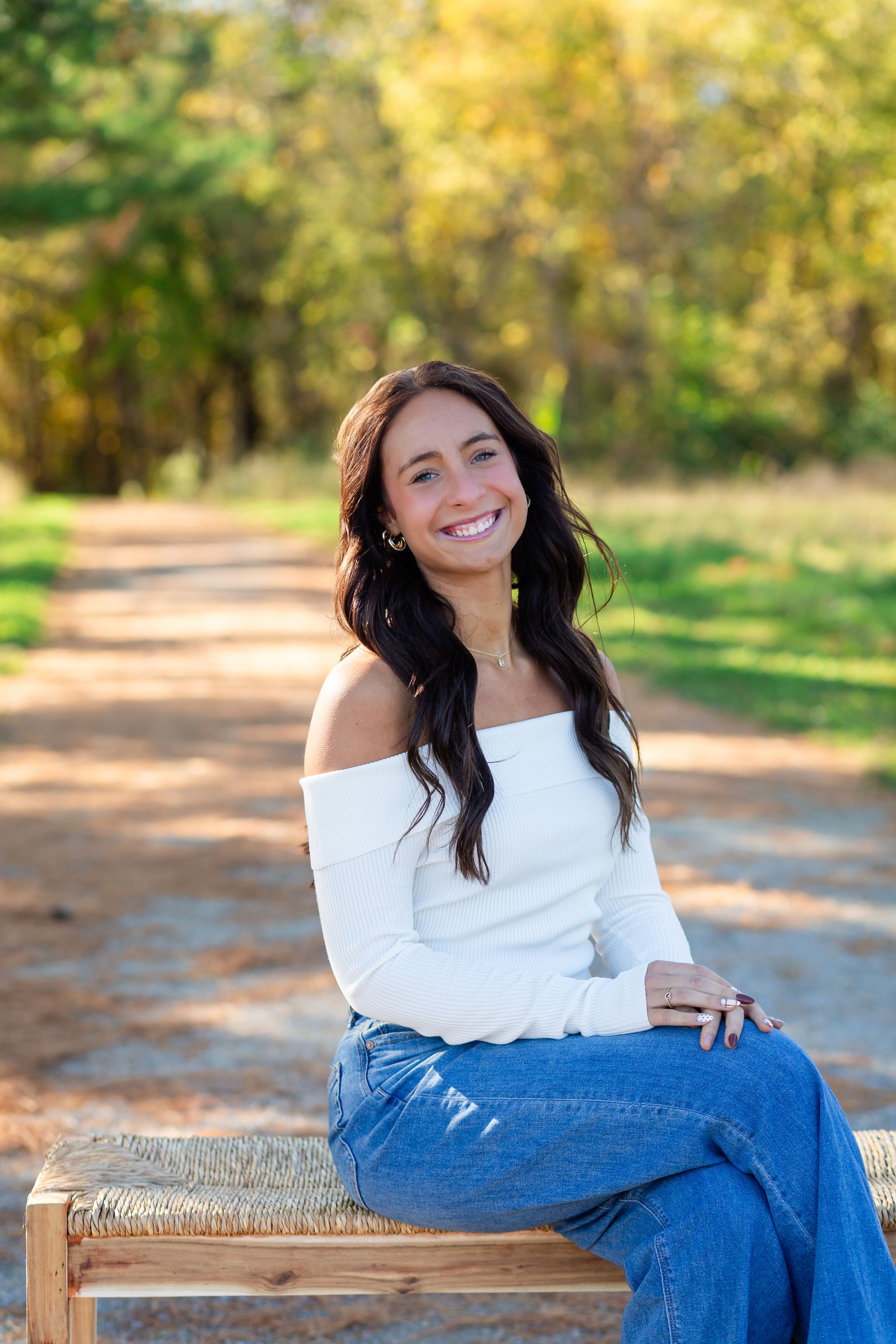 A person in white off-shoulder top and jeans poses casually on a hay bale outdoors during autumn.