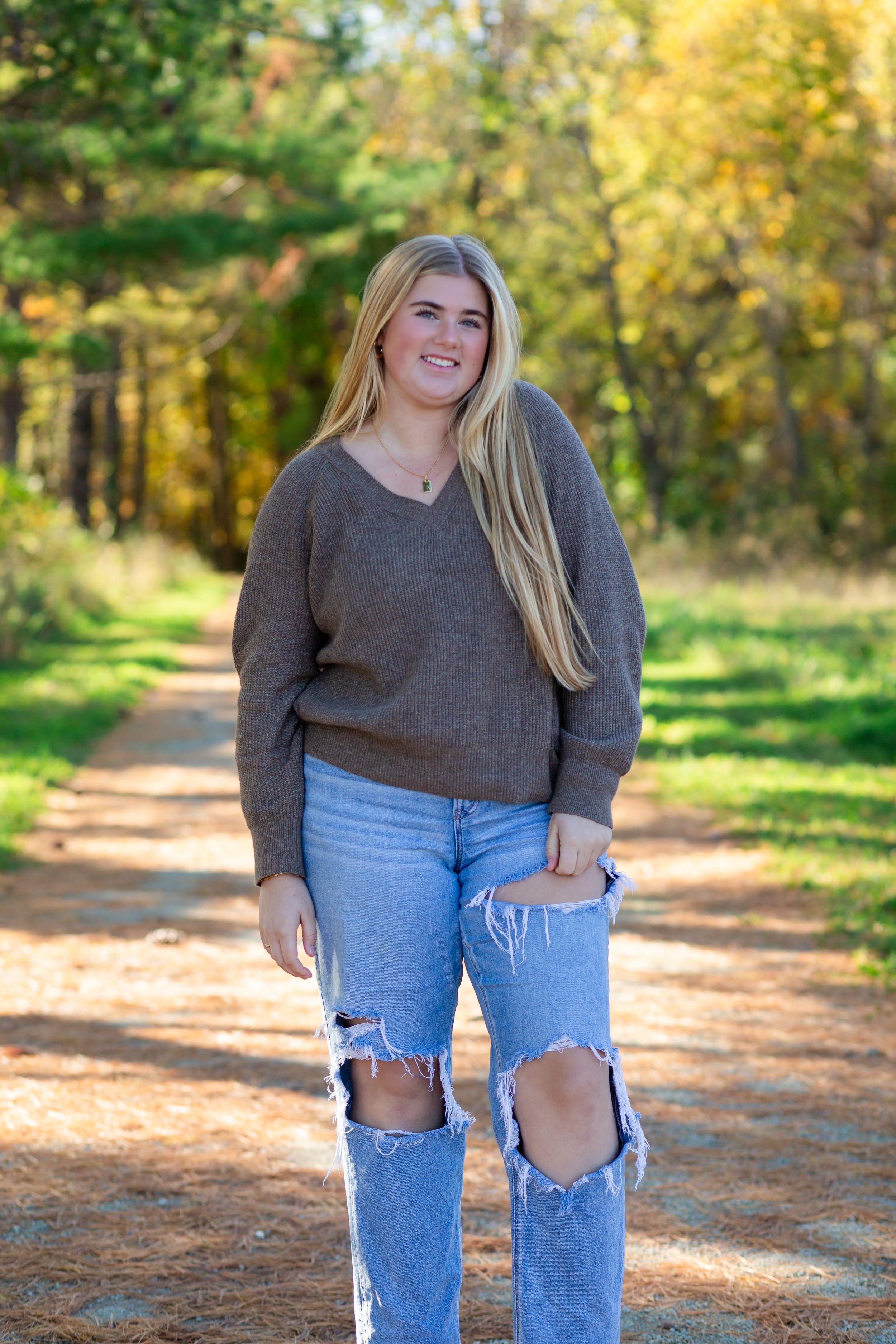 A person in a gray sweater and distressed jeans stands on a dirt path surrounded by autumn foliage.