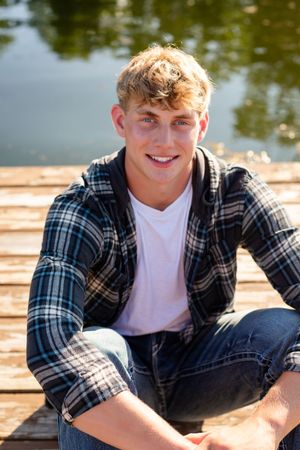 A person in a plaid flannel shirt and white t-shirt sits outdoors on a sunny day near water.