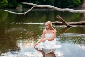 A person in a white wedding dress poses gracefully on a fallen tree trunk over a tranquil lake.
