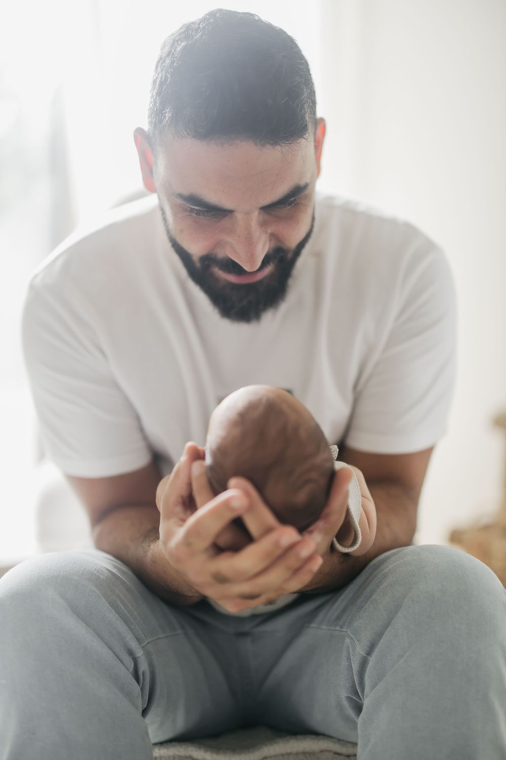 A bearded parent in a white t-shirt tenderly holds and gazes at their newborn baby while sitting down.