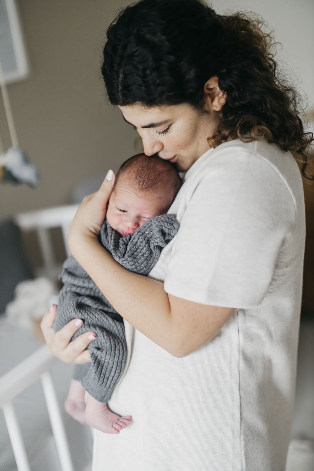 A parent in white clothing cradles and kisses a sleeping newborn baby wrapped in a gray blanket.