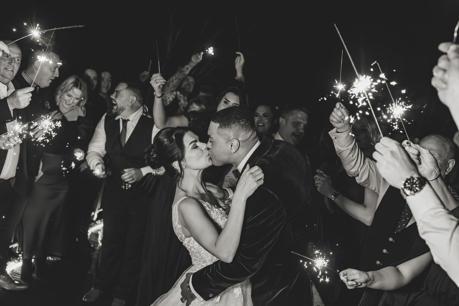 Wedding party with sparklers as bride and groom share a kiss.