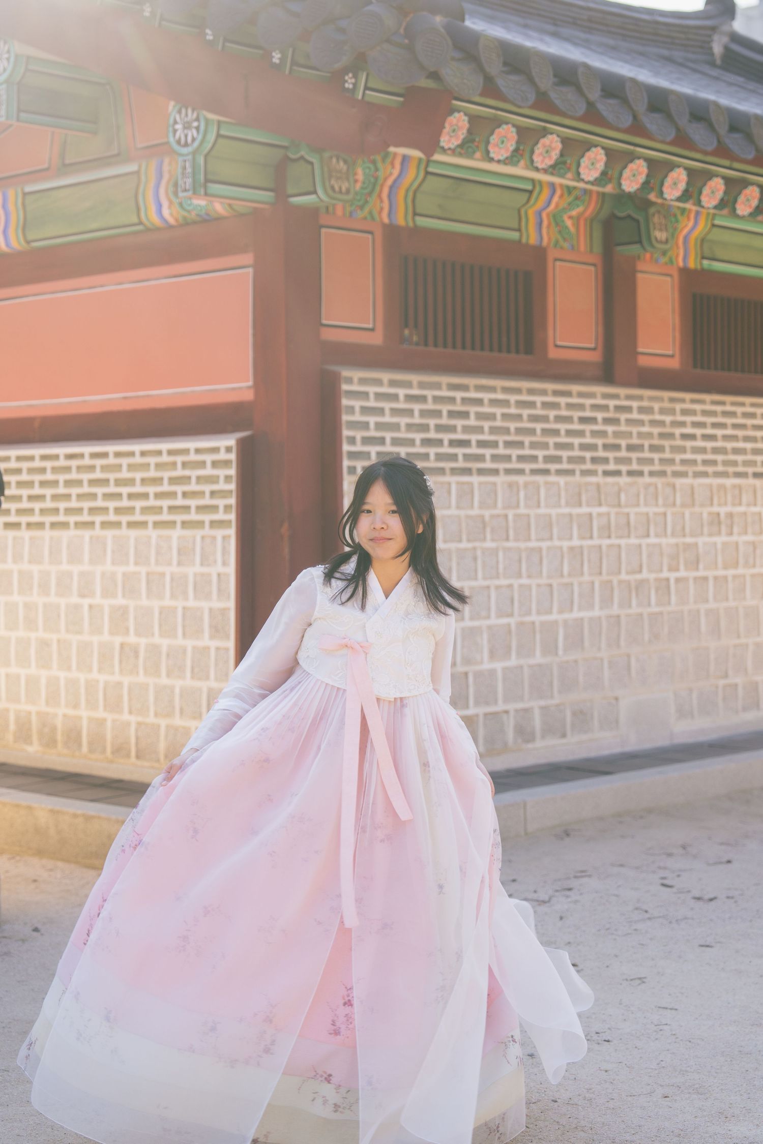 A figure in a flowing pink and white traditional hanbok dress twirls gracefully in front of a Korean palace wall.