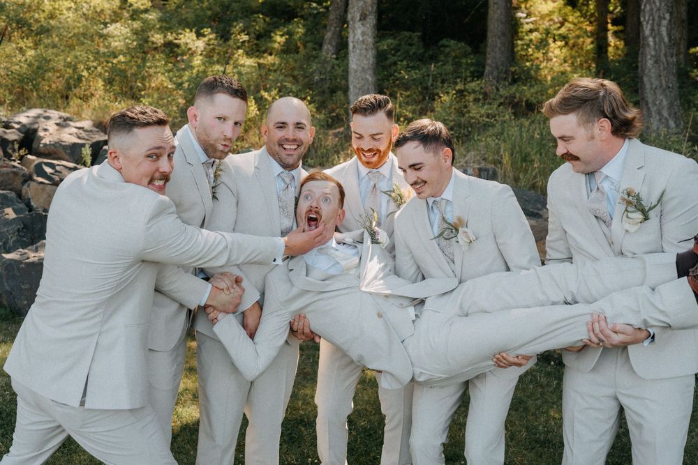 Wedding party in light gray suits laughing and celebrating together outdoors in a natural setting.