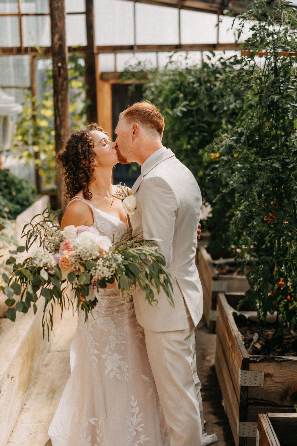A romantic wedding couple sharing a kiss in a greenhouse setting with a lush floral bouquet.