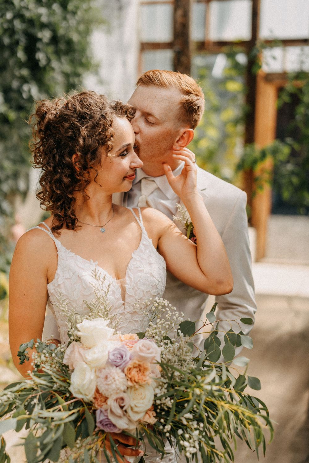 A romantic wedding couple shares intimate moments while holding a pastel floral bouquet with eucalyptus in an outdoor setting.