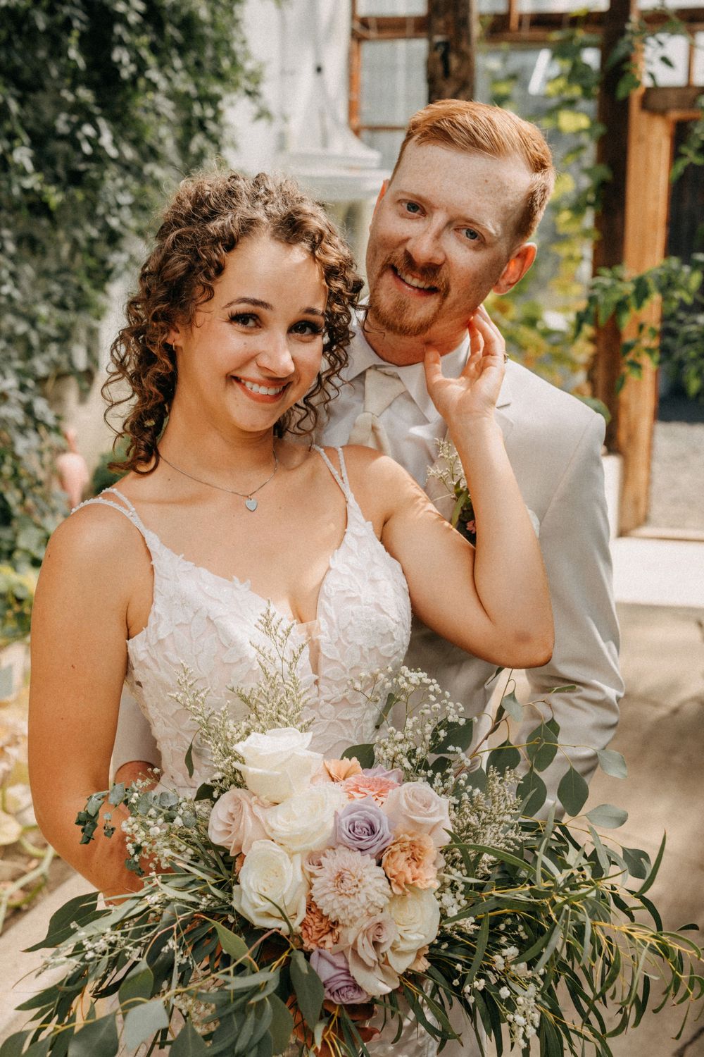 A joyful wedding portrait in a greenhouse with a beautiful bouquet of mixed flowers.