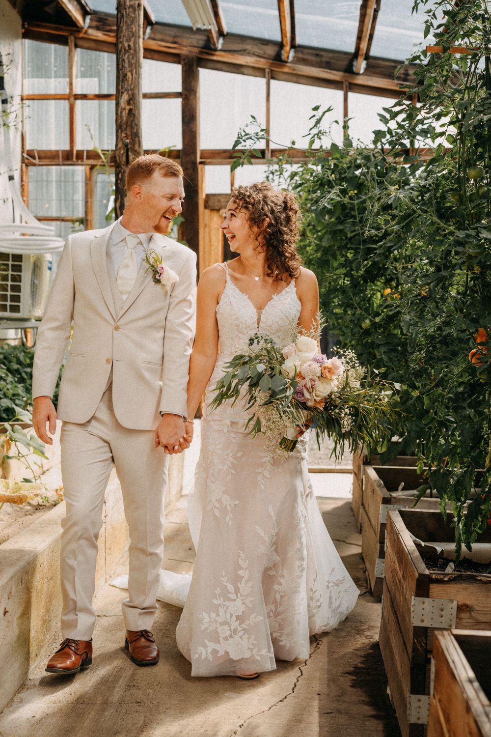 A wedding couple walking hand in hand through a rustic greenhouse pathway.