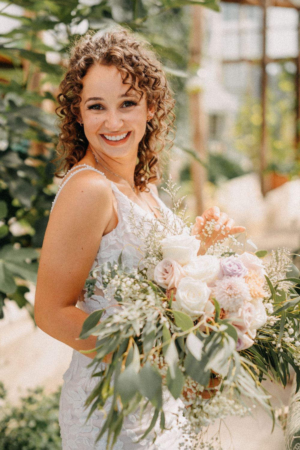 A radiant bride holding a lush bouquet of pink roses and eucalyptus in a greenhouse setting.