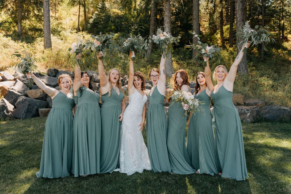 Wedding party in sage green dresses poses together outdoors in a sunlit garden setting.