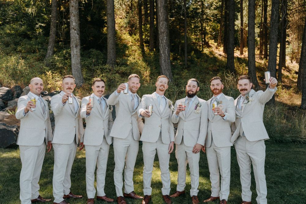 A large wedding party in white attire poses together outdoors in a natural wooded setting.