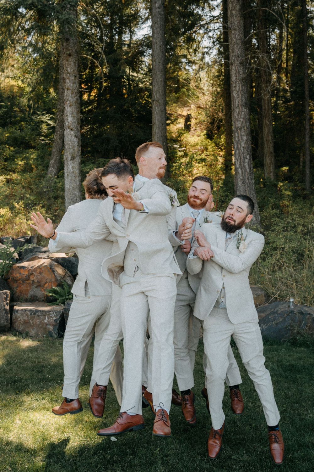 Groomsmen in cream colored suits playfully pose together in a forest setting.