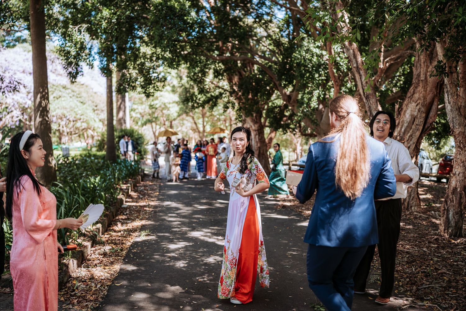 Wedding guests gather on a tree-lined path during an outdoor celebration.