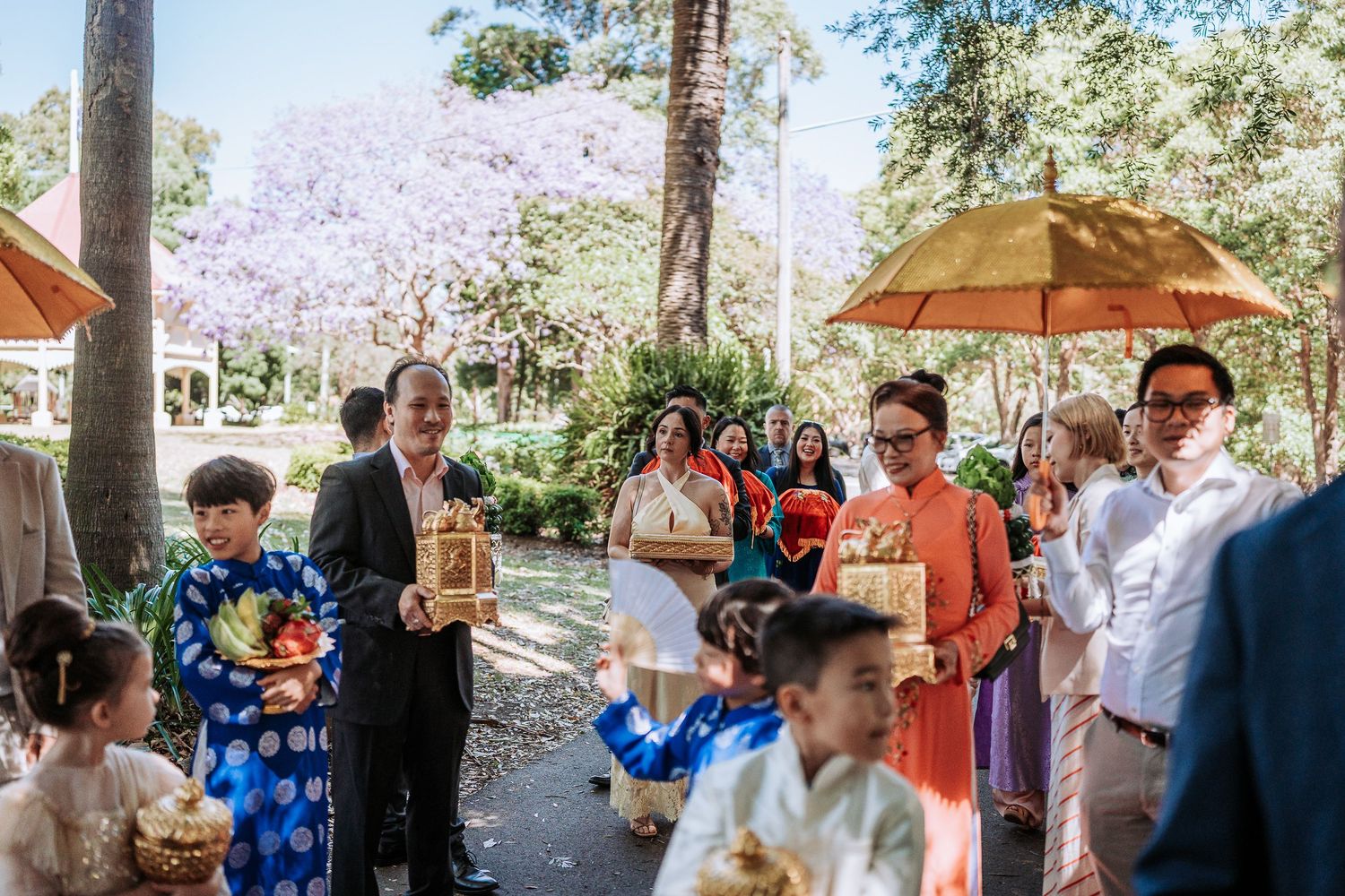 Outdoor wedding ceremony in a garden setting with guests seated under trees and natural sunlight filtering through.