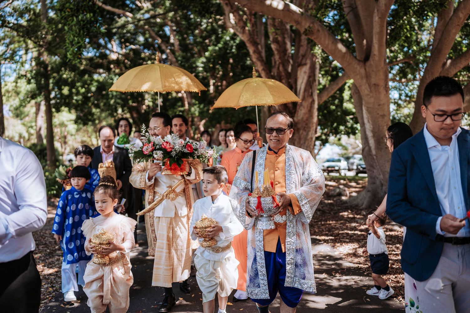 Wedding guests gather outdoors under trees for a celebration with drinks and umbrellas.