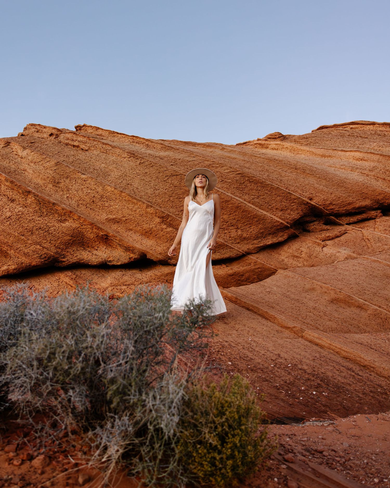 Figure in white dress stands against dramatic red rock formations in a desert landscape at sunset.
