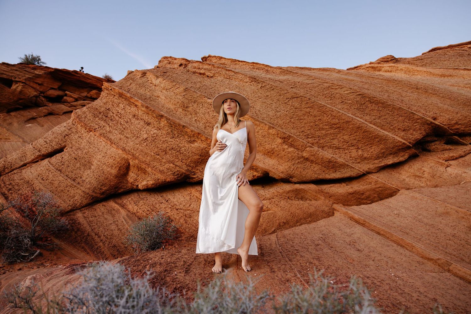 A person in a flowing white dress poses gracefully against the backdrop of red desert rock formations at sunset.