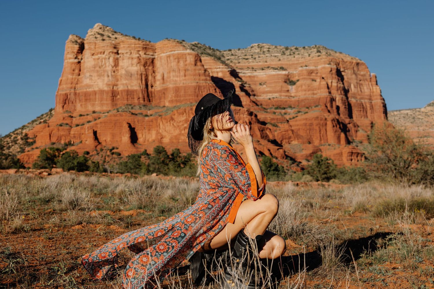 A scenic view of red rock formations in Sedona with someone in a patterned dress sitting on a rock formation.