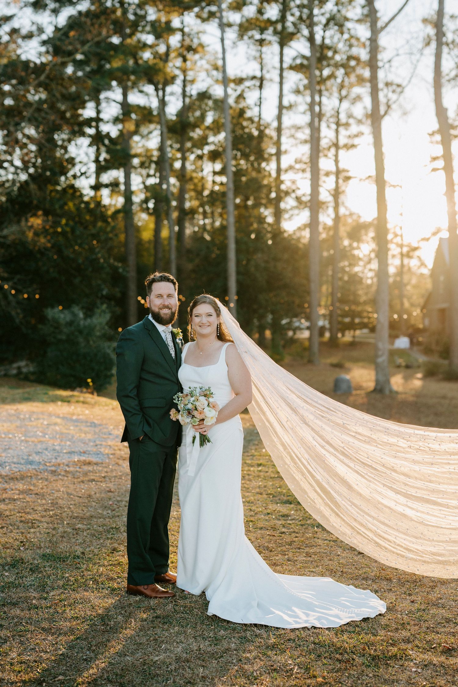 A romantic sunset wedding portrait in a pine forest with a flowing cathedral veil catching the golden light.