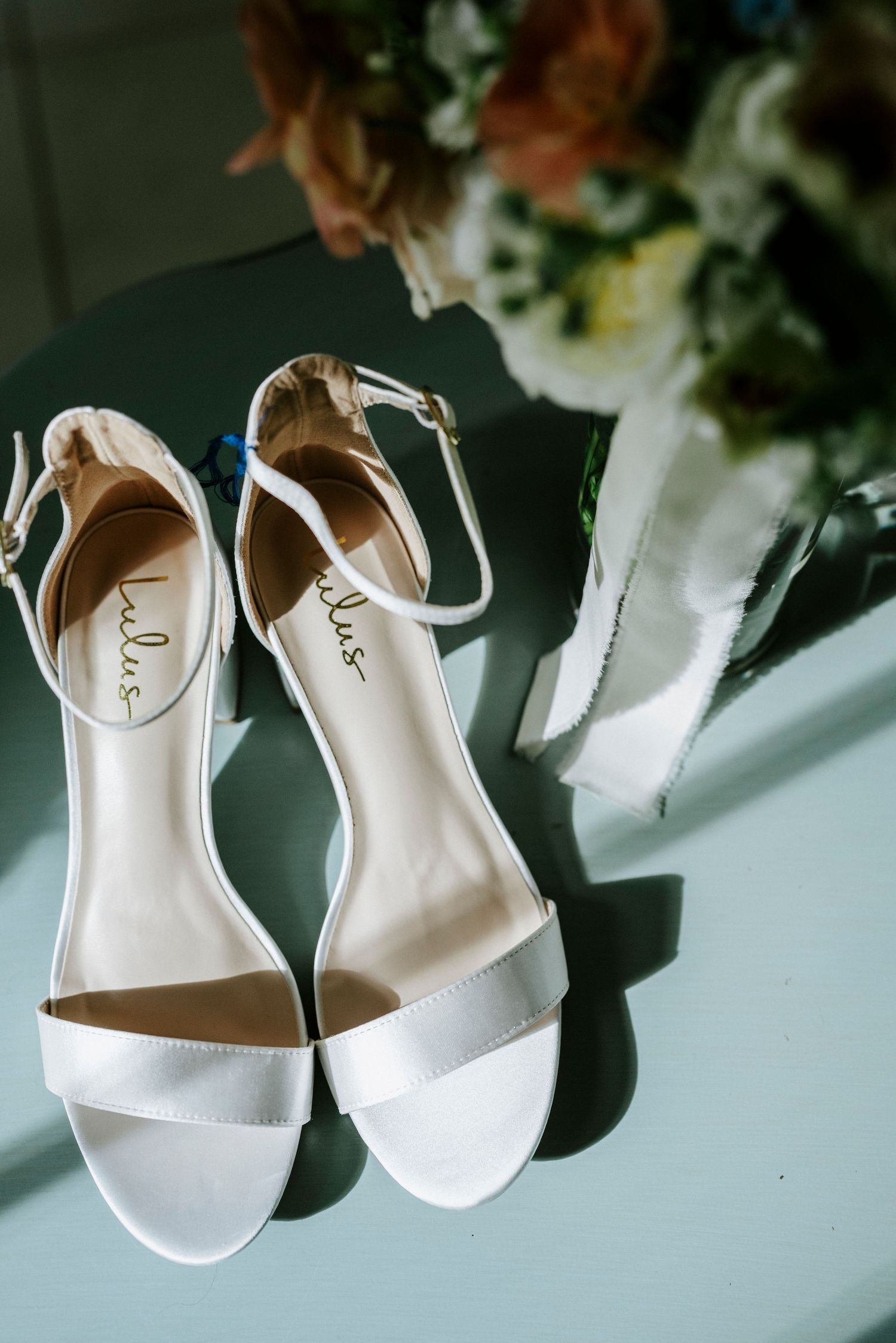 Elegant white bridal sandals with ankle straps photographed in natural light with soft floral shadows.