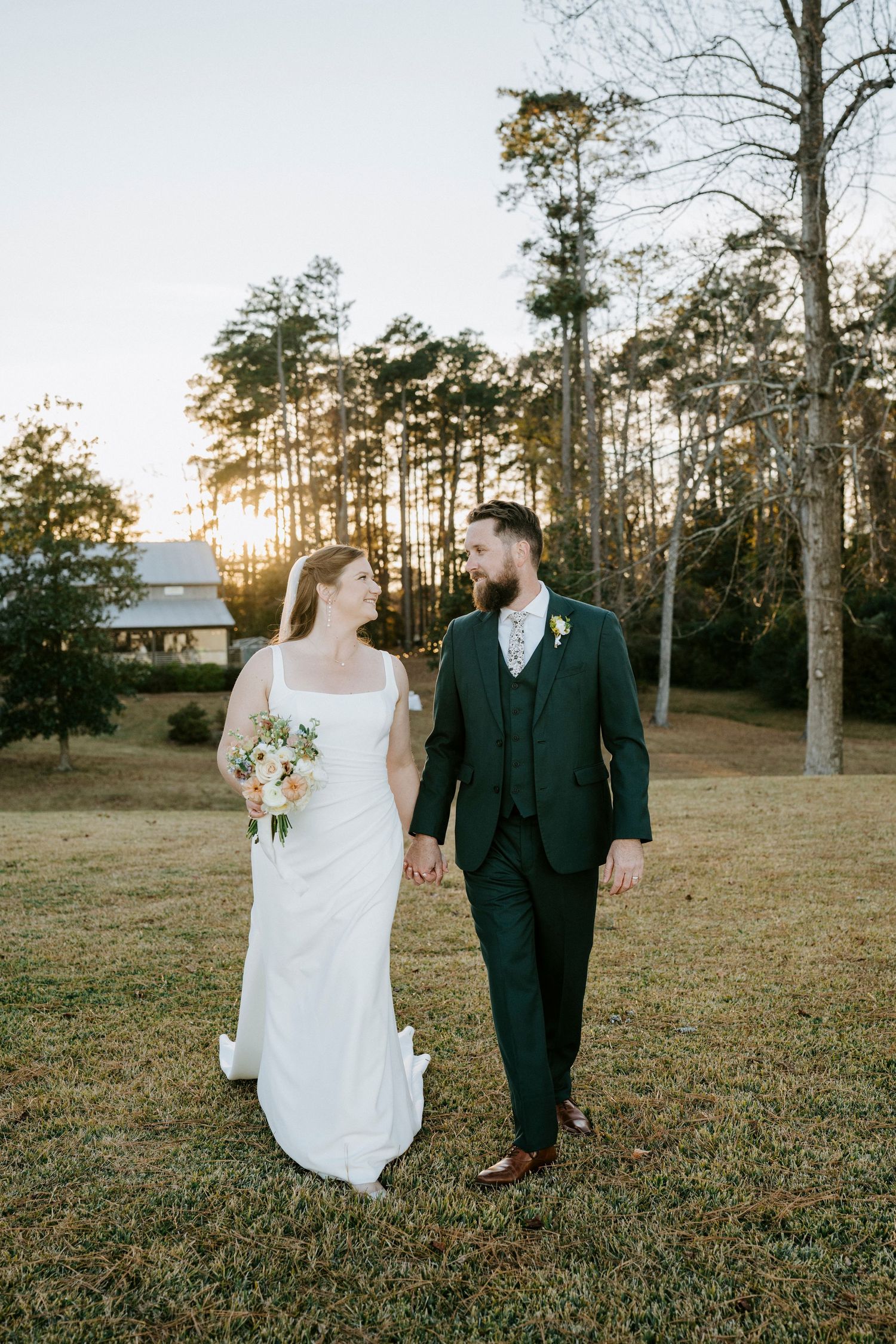 A bride and groom walk together through a sunlit field with tall pine trees in the background during golden hour.